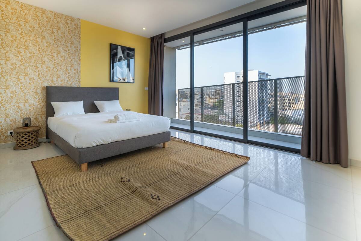 A modern hotel room with a large bed, a view of the city through floor-to-ceiling windows, and a yellow accent wall.