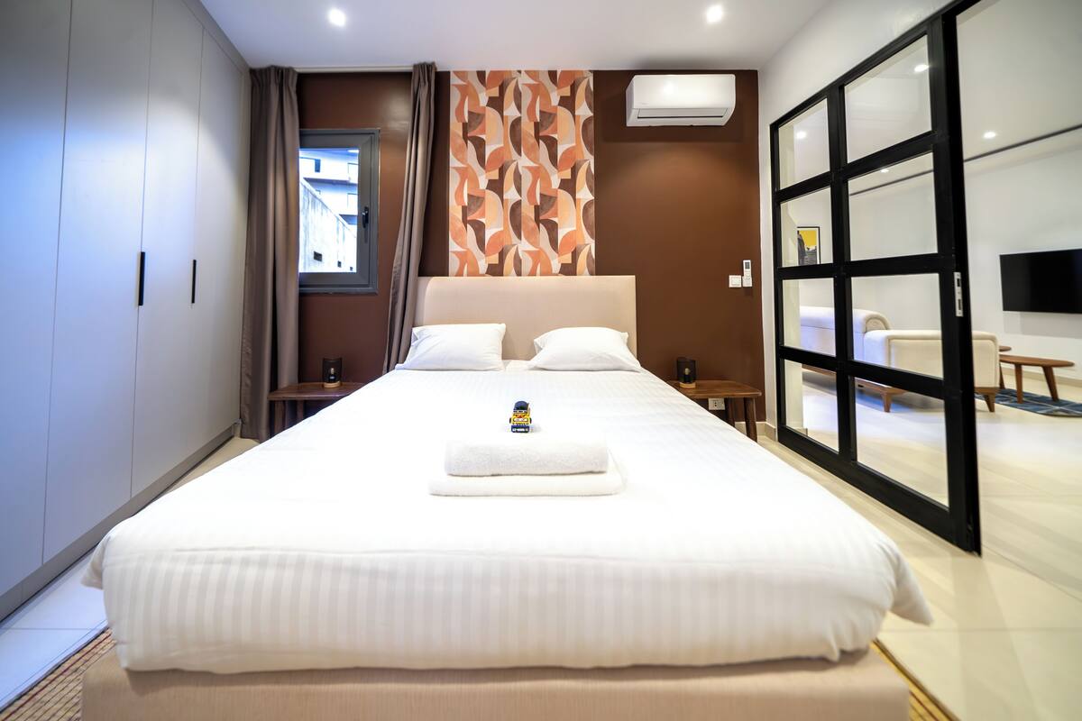 A hotel room with a large bed, a flat-screen TV, a window with curtains, and a wall-mounted air conditioner.