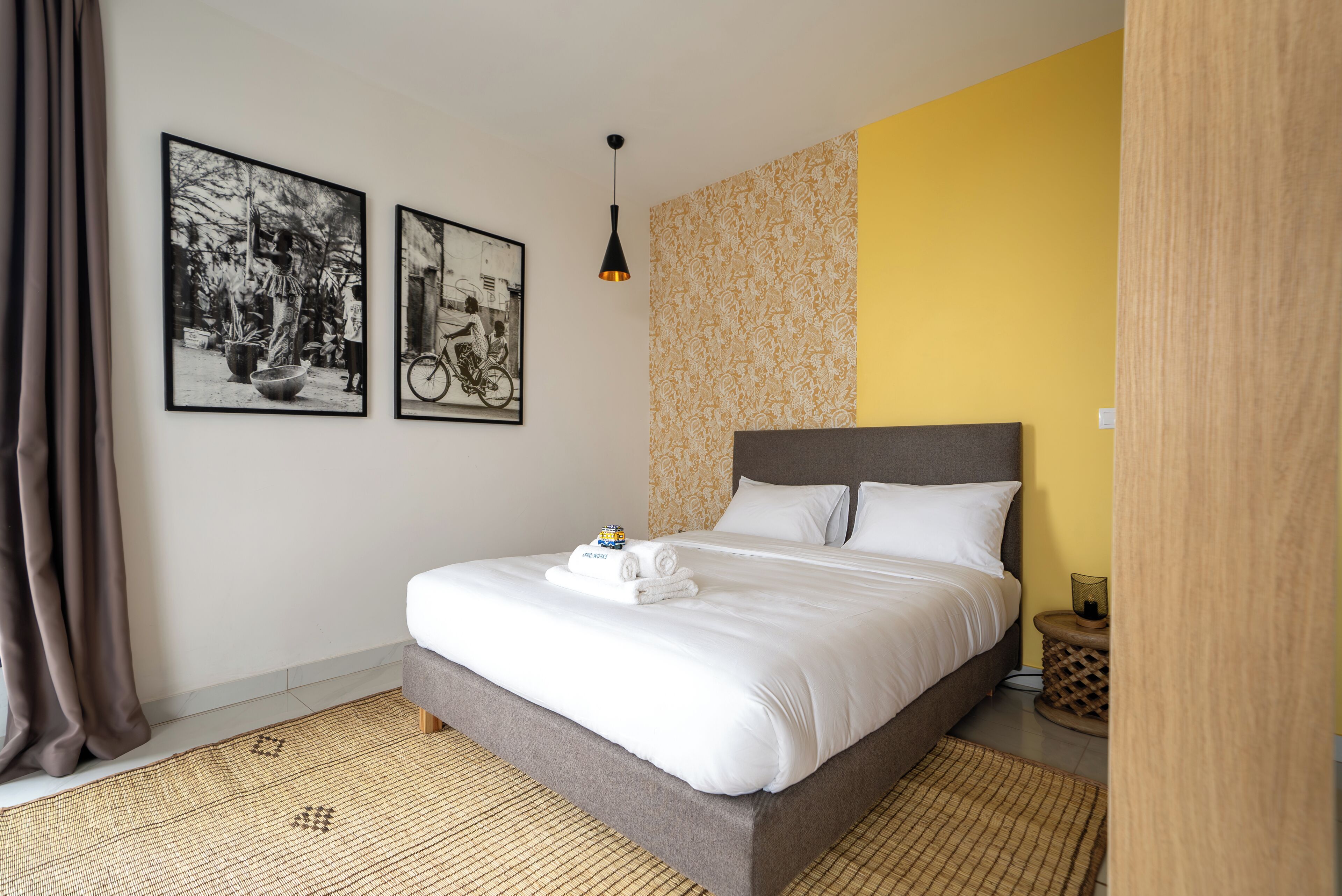 Un lit bien fait, avec une tête de lit grise et du linge de lit blanc, une petite table de chevet avec une lampe, et deux tableaux encadrés accrochés au mur.