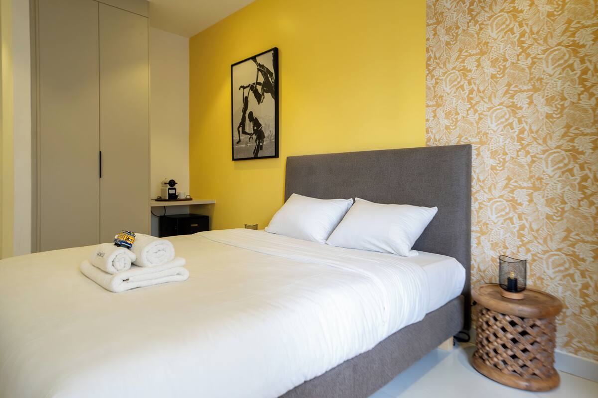 A neatly made bed with white linens and rolled towels, a bedside table with a lamp, and a framed picture on the wall.