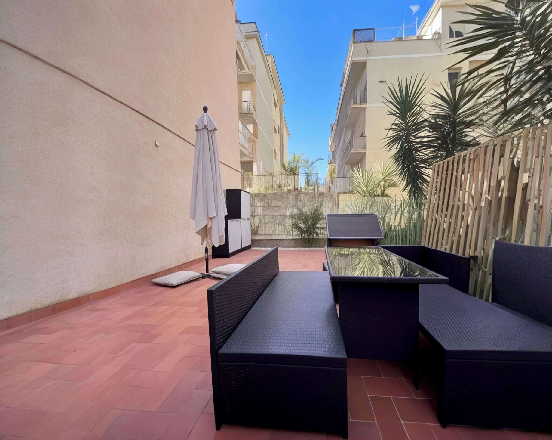 Eine Terrasse im Freien mit einem schwarzen Tisch und zwei schwarzen Sofas, einem weißen Sonnenschirm und einem Holzzaun.