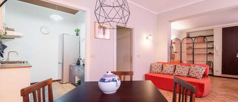 Un comedor con mesa de madera, sillas, un cuenco decorativo, un sofá rojo con cojines estampados y una lámpara colgante de diseño geométrico.