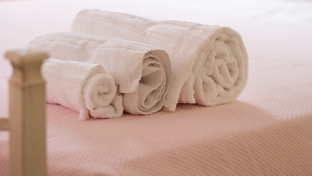 Una cama bien tendida con toallas blancas enrolladas, un cabecero de madera, una mesita de noche con lámpara y una ventana al fondo.