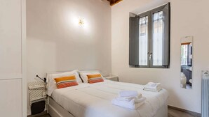Un dormitorio con cama, almohadas, mesita de noche, luz adosada a la pared, ventana y radiador.
