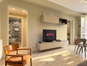 Uma sala de estar moderna com uma televisão de tela plana exibindo 'NETFLIX', um móvel branco para equipamentos eletrônicos e uma cadeira de madeira.