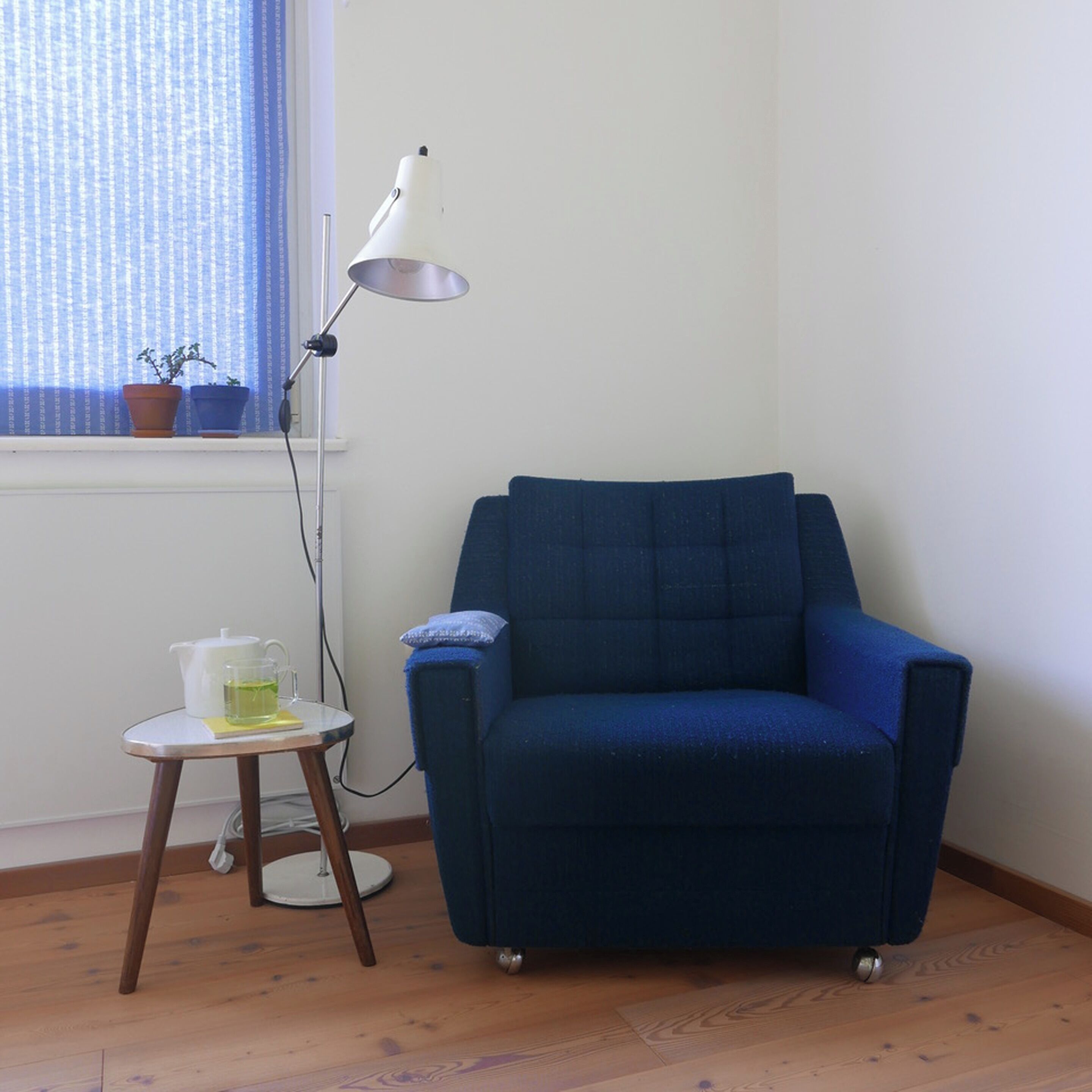 Ein Zimmer mit blauem Sessel, einem kleinen Tisch mit Teekanne und Tasse sowie einer Stehlampe.