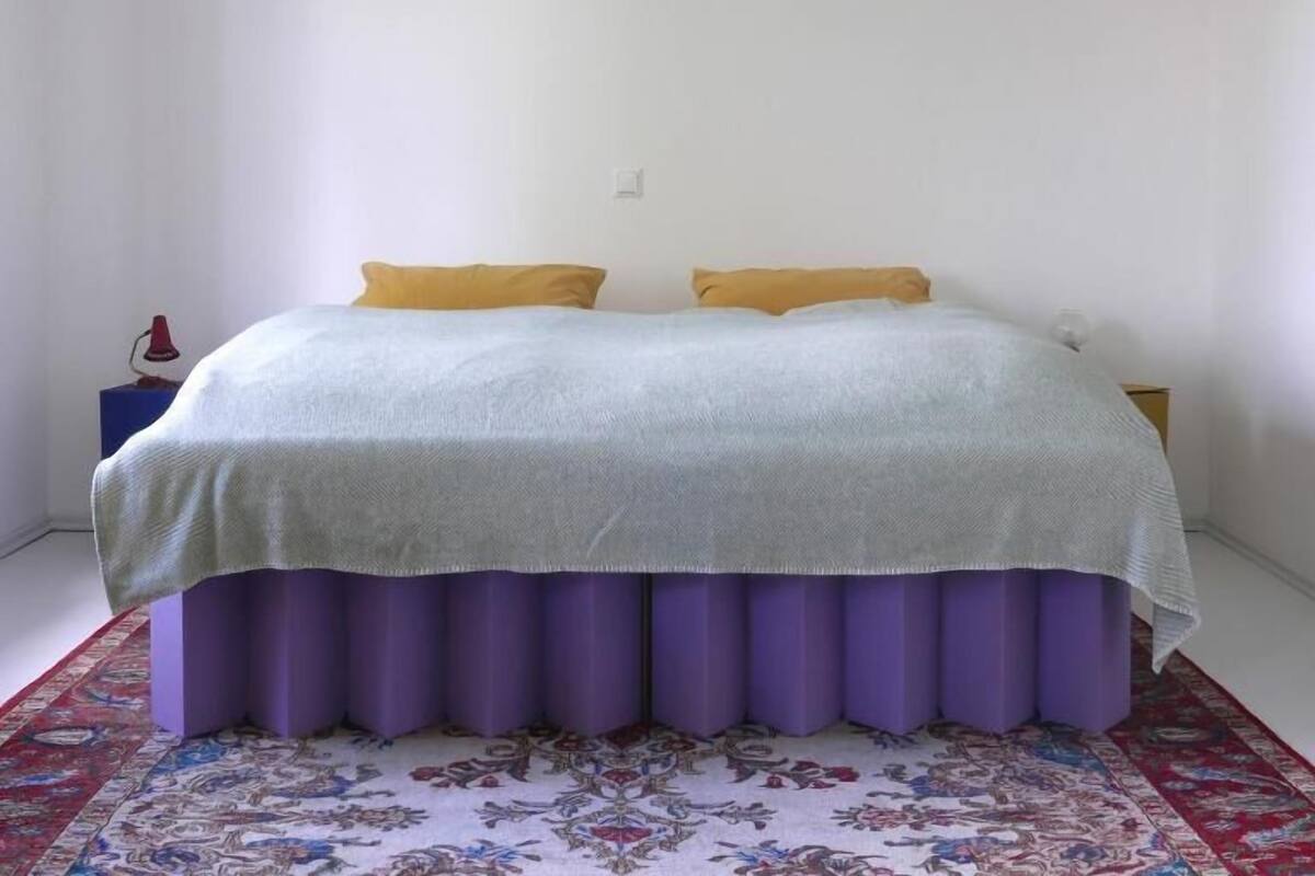 Ein Schlafzimmer mit einem Bett, zwei Kissen, einem Nachttisch, einer Lampe und einem gemusterten Teppich.