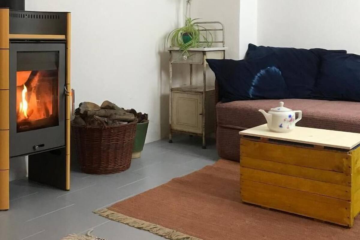 Ein gemütliches Wohnzimmer mit Kaminofen, einer Couch, einem kleinen Tisch mit Teekanne und einer Lampe.
