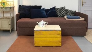 A living room with a brown sofa, a yellow wooden chest, and a white teapot on a small table. - Unter einem Dach (Raabs An Der Thaya)