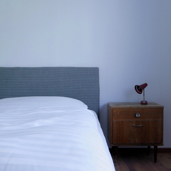 Una cama con sábana blanca, una mesita de noche de madera con una lámpara roja y un cabecero de ladrillo gris.