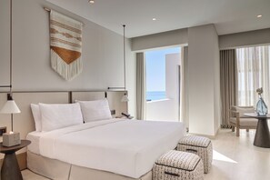 Una habitación de hotel moderna con una cama grande, dos mesitas de noche, una zona de estar con un sillón y reposapiés, un tapiz colgado en la pared, y vistas al océano a través de amplios ventanales.