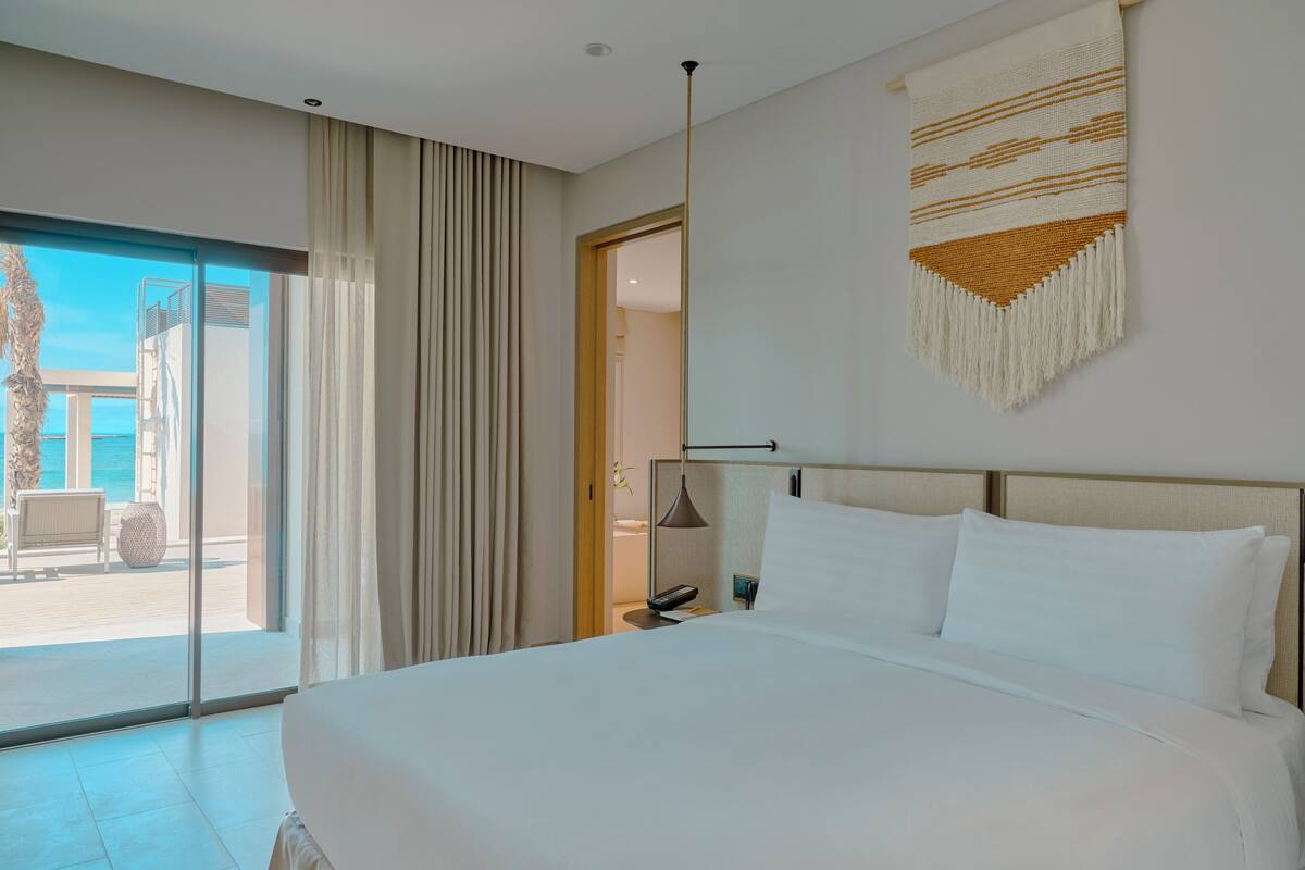 A modern hotel room with a large bed, a view of the beach, and a balcony with a chair and table.