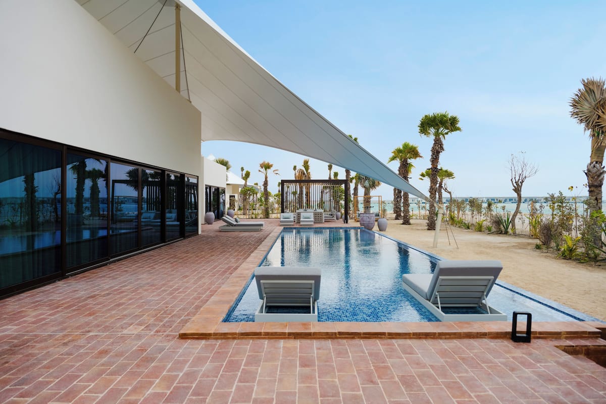 A modern outdoor pool area with a clear view of the beach, palm trees, and a white building with large windows.