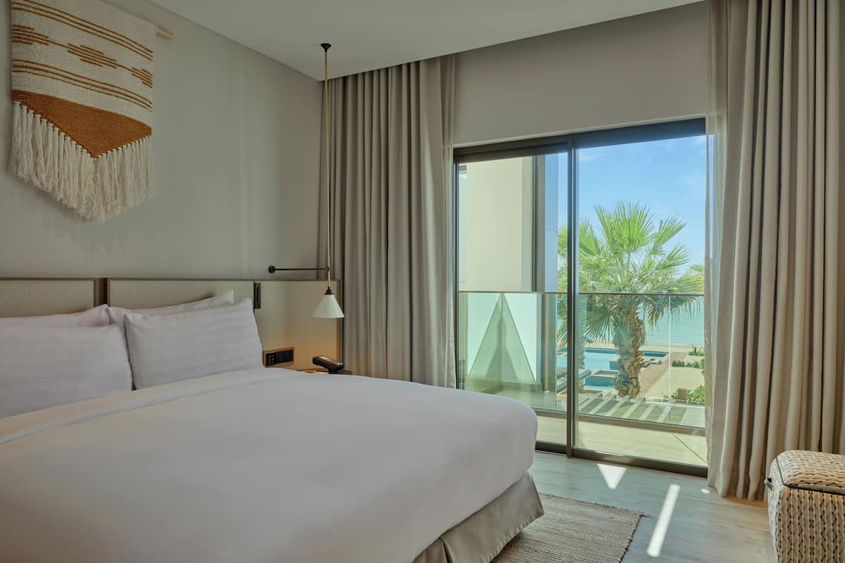 A modern hotel room with a large bed, a view of a pool and palm trees, and a balcony with curtains.