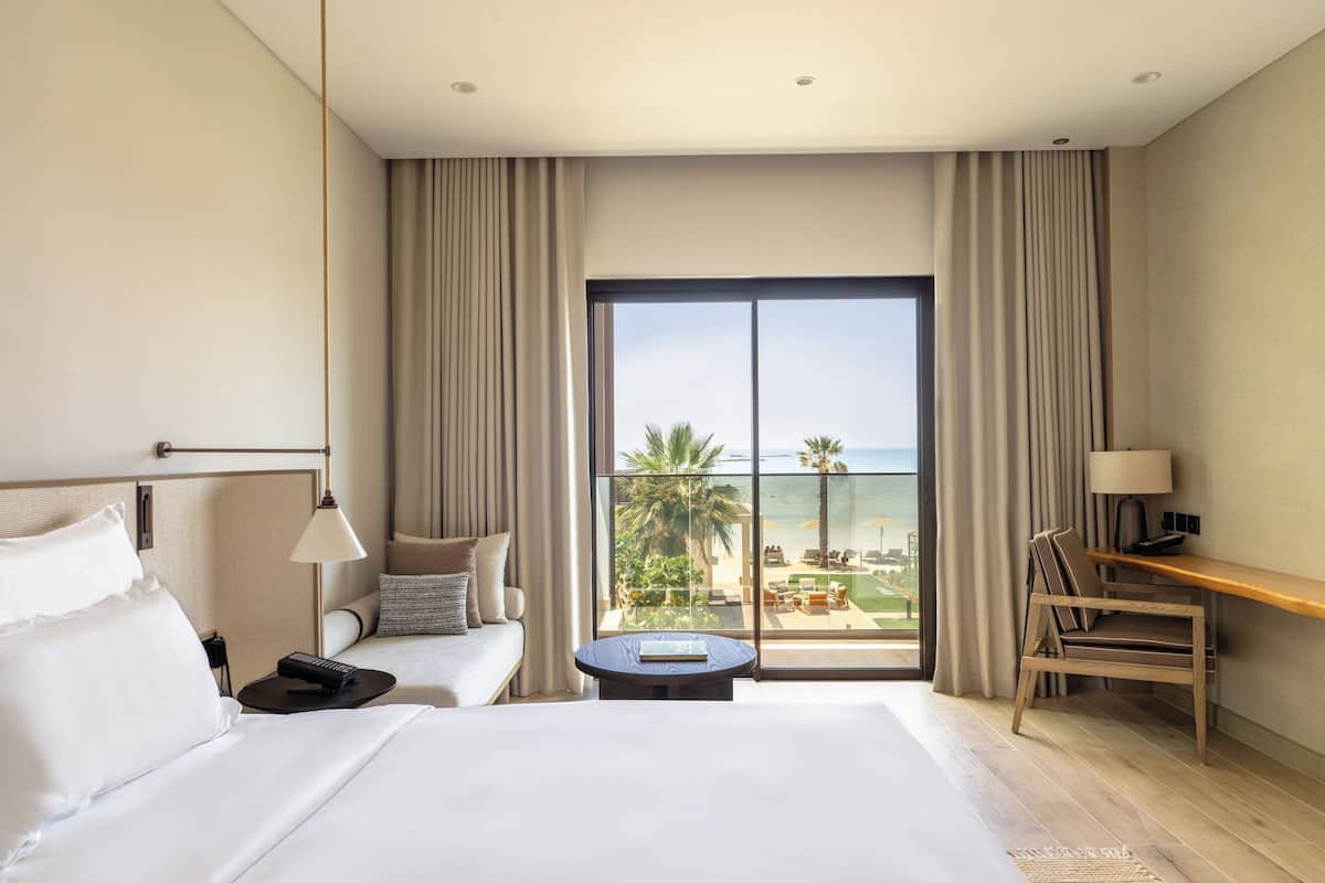 A hotel room with a large bed, a desk, a chair, a round table, and a view of the beach and palm trees through a large window.