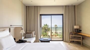 A hotel room with a large bed, a desk, a chair, a round table, and a view of the beach and palm trees through a large window.