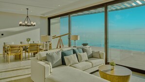 A spacious living area with a large window overlooking the ocean, a dining set, and a modern chandelier. - Hawar Resort By Mantis (Hawar)