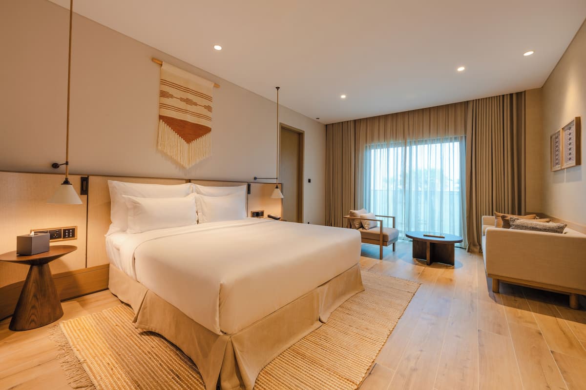A modern hotel room with a large bed, a sitting area with a sofa and a small table, and a large window with curtains.