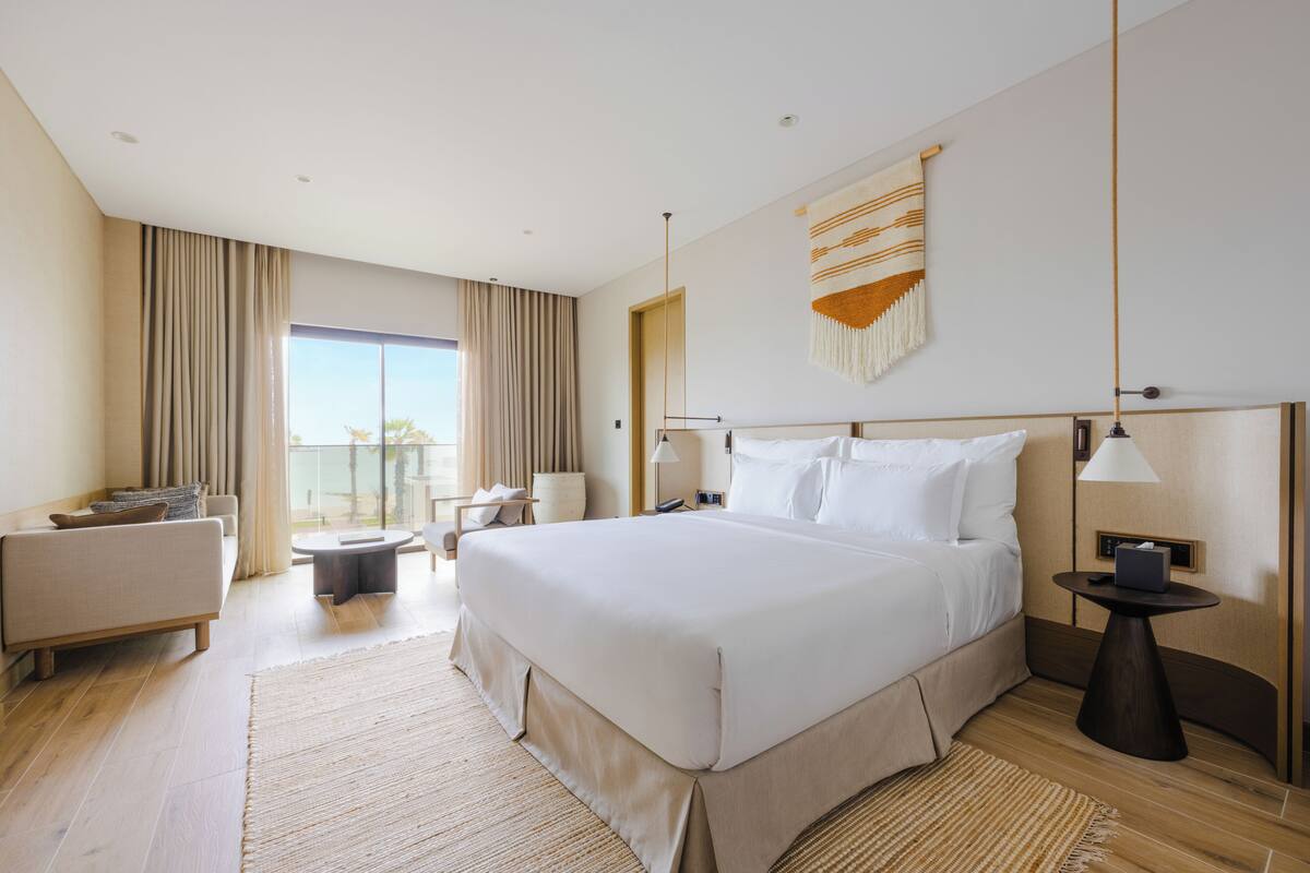 A modern hotel room with a large bed, a sofa, a small table, and a view of the outdoors.