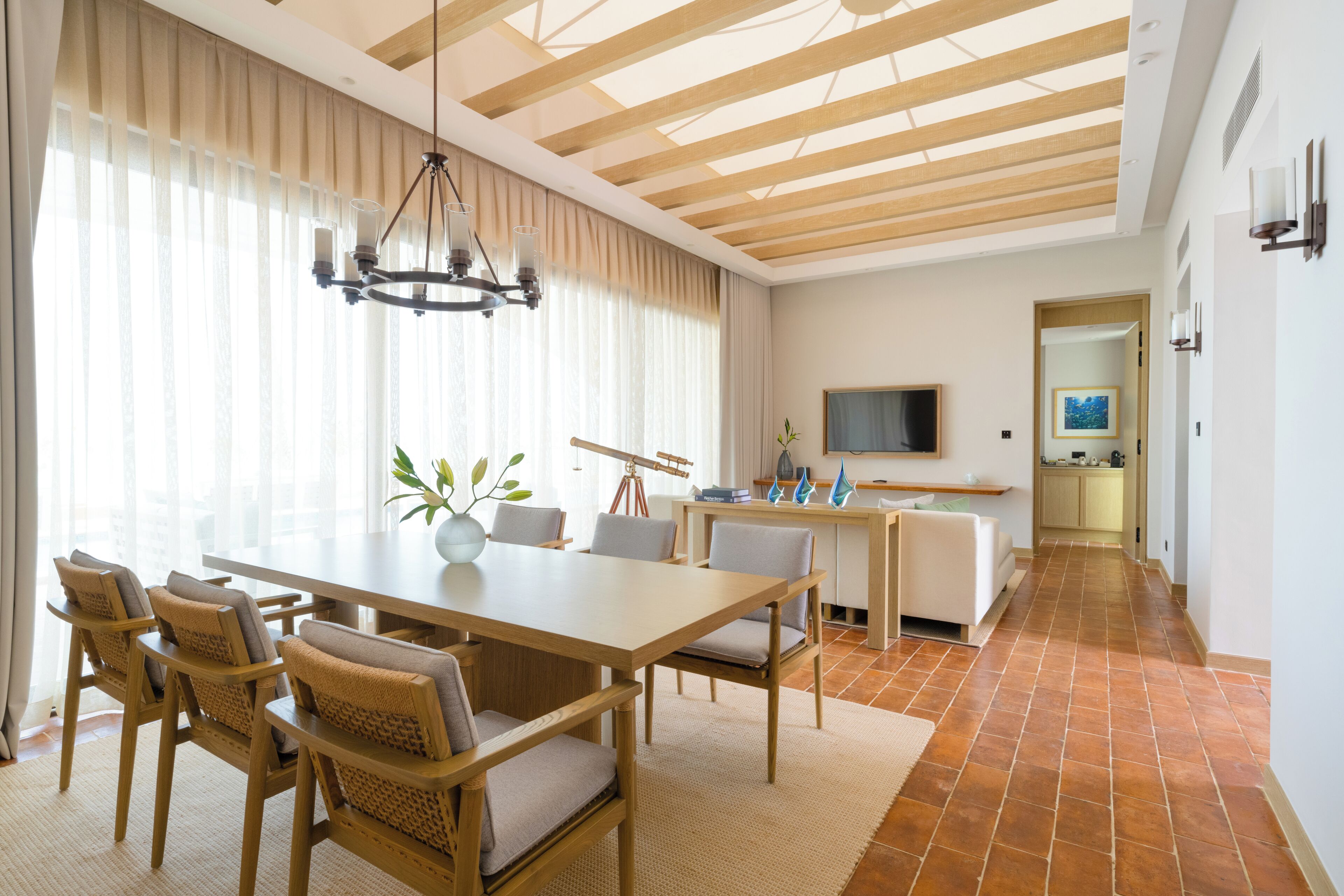 Un comedor amplio con una mesa y sillas de madera, una araña y vistas a la cocina al fondo.