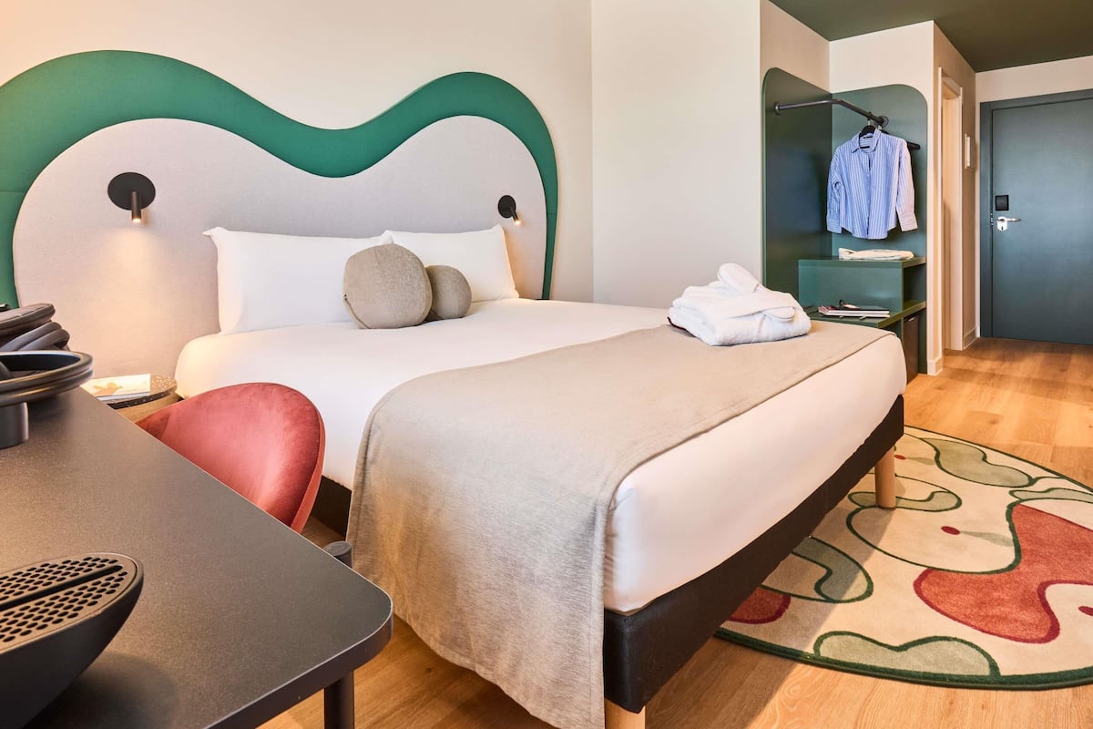 Une chambre d’hôtel avec un lit, une table de chevet, une chaise et une salle de bain visible à travers une porte ouverte.