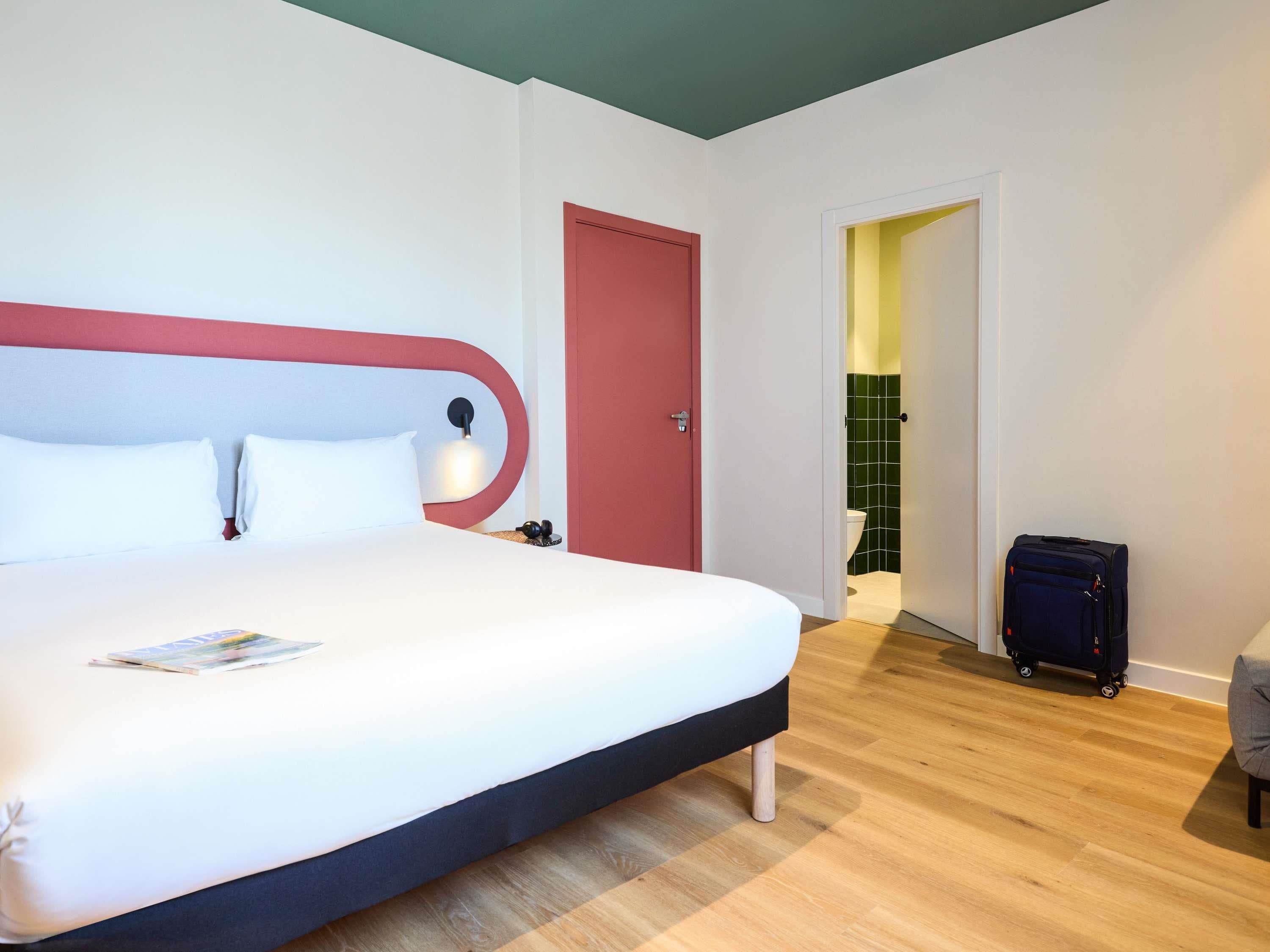 Una habitación de hotel con una cama, una mesita de noche, una maleta y un baño visible a través de una puerta abierta.