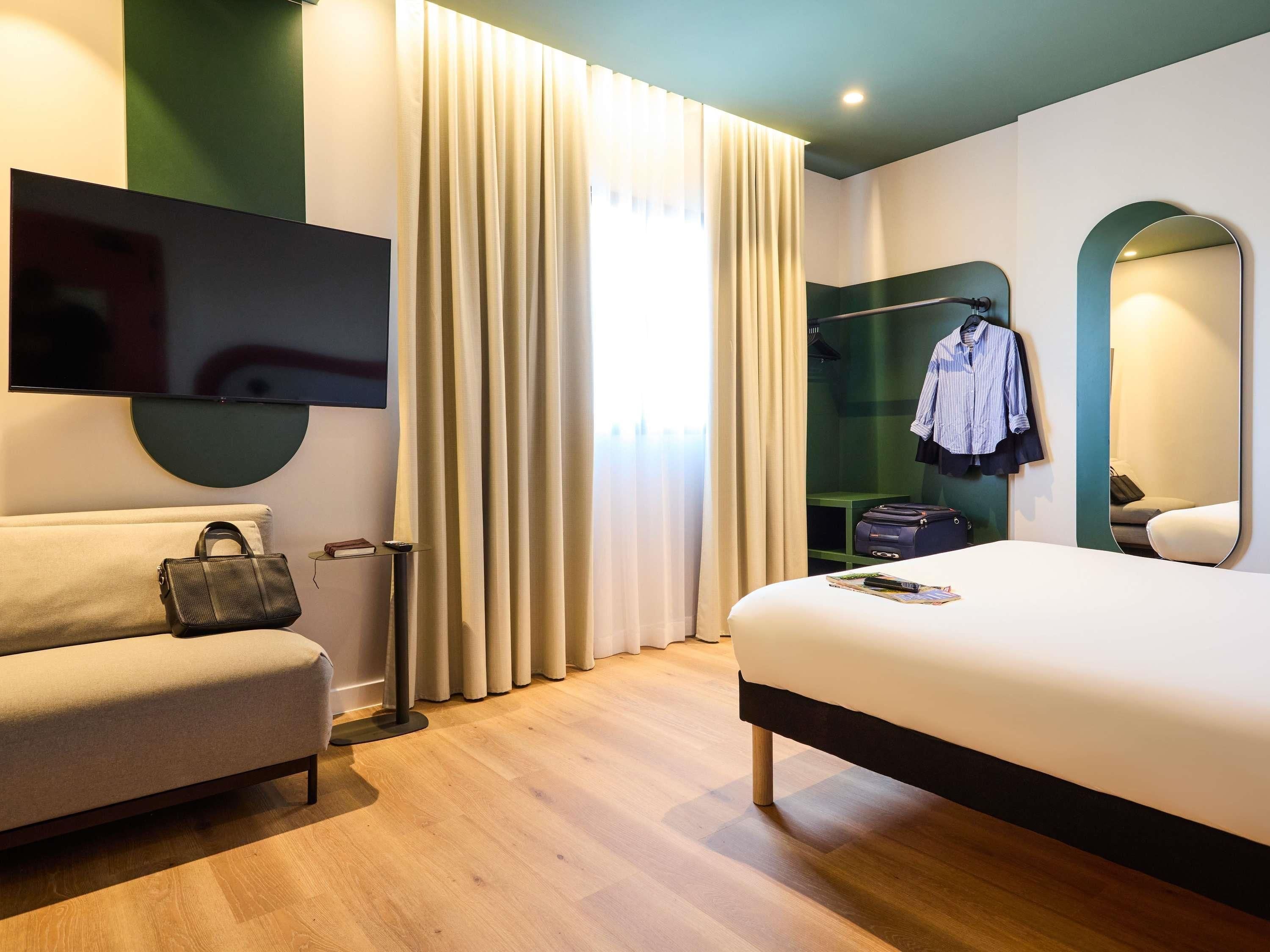 A modern hotel room with a bed, a sofa, a TV, a mirror, and a wardrobe.