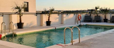 Una piscina en la azotea con vistas despejadas del paisaje urbano, rodeada de macetas con plantas y sillones de descanso.