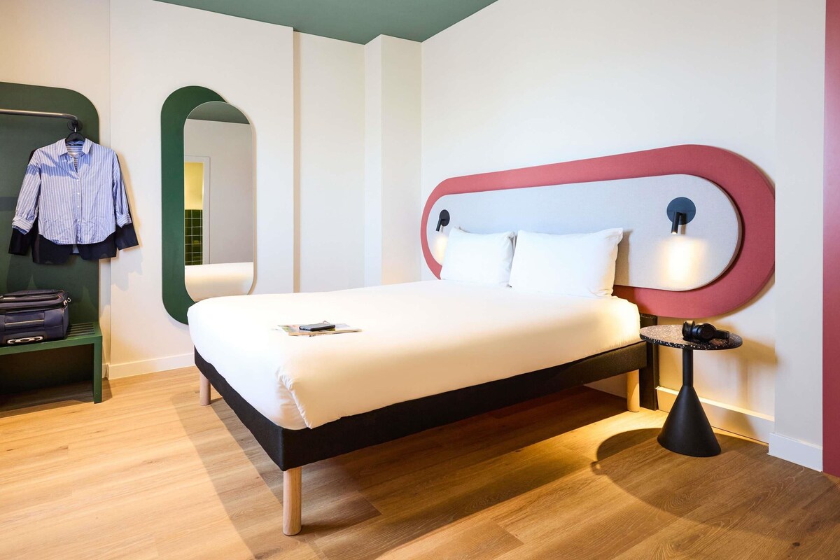 Une chambre d’hôtel moderne équipée d’un lit, d’une table de chevet, d’une lampe, d’un miroir et d’une armoire.