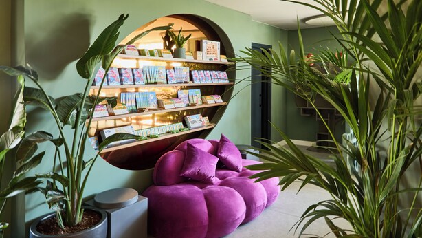 Een kamer met een ronde wandplank waarop boeken en een potplant staan, een paarse kruikzak en een grote potplant.