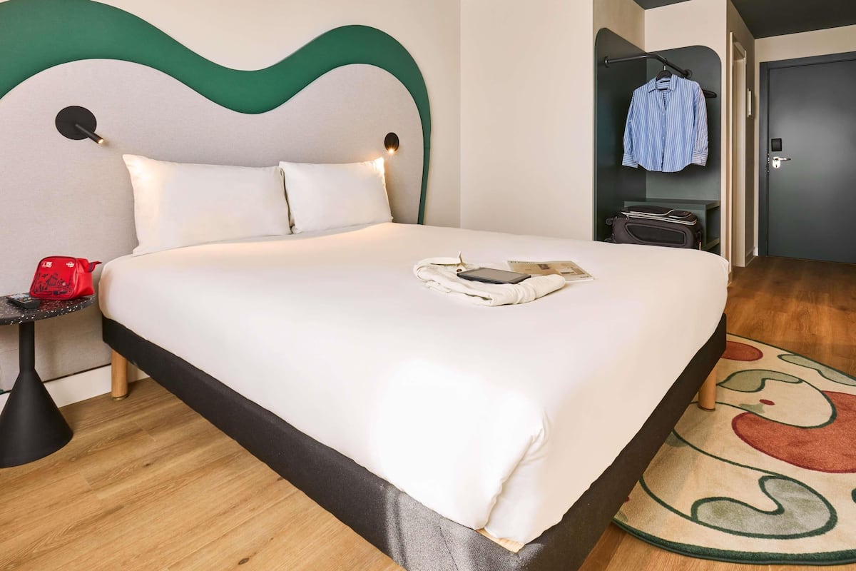 Een hotelkamer met een bed, een nachtkastje met een rode tas, een kapstok met een gestreept shirt en een koffer.