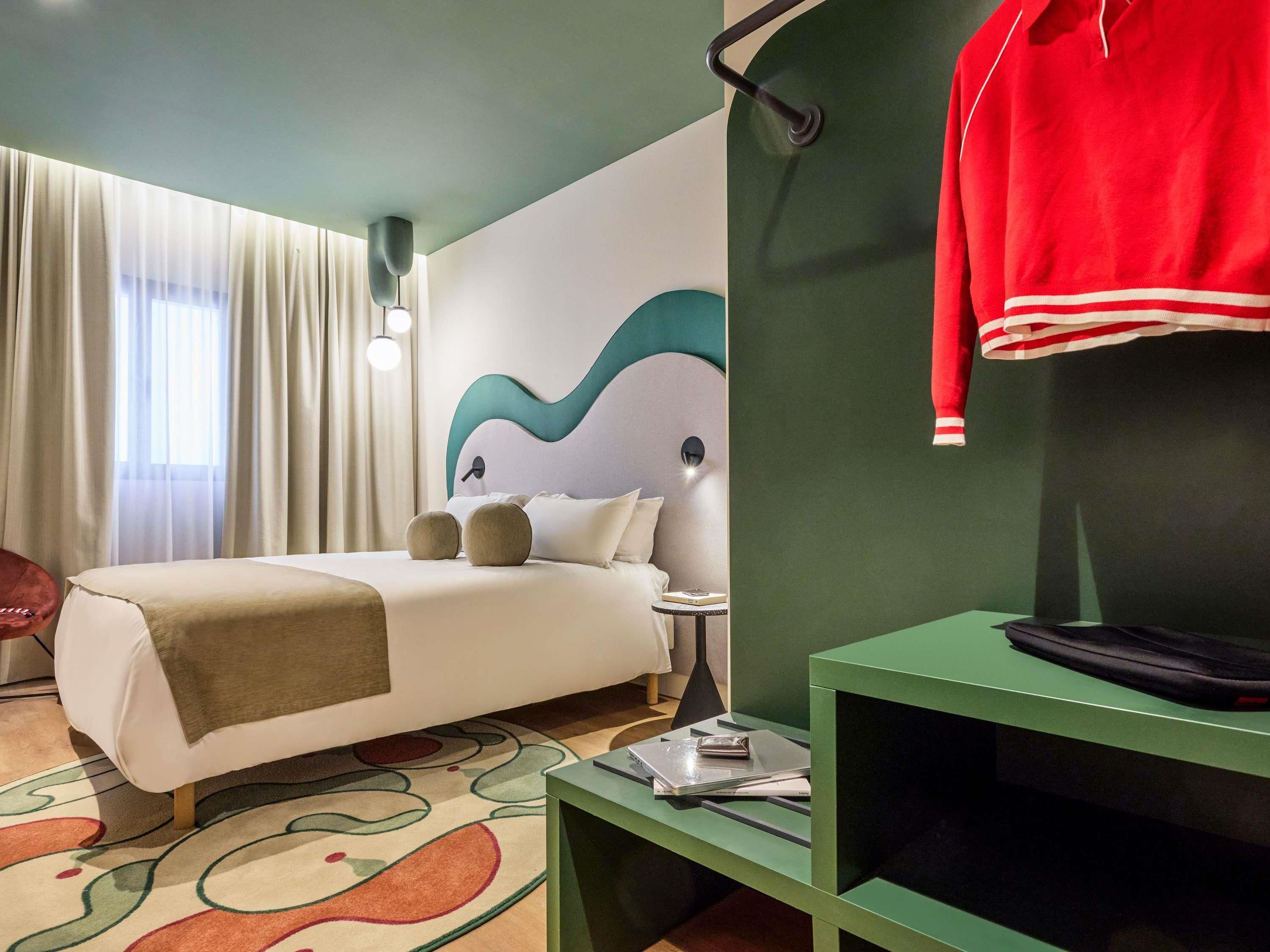 A hotel room with a bed, a green side table with a laptop, a red jacket hanging on the wall, and a large window with curtains.