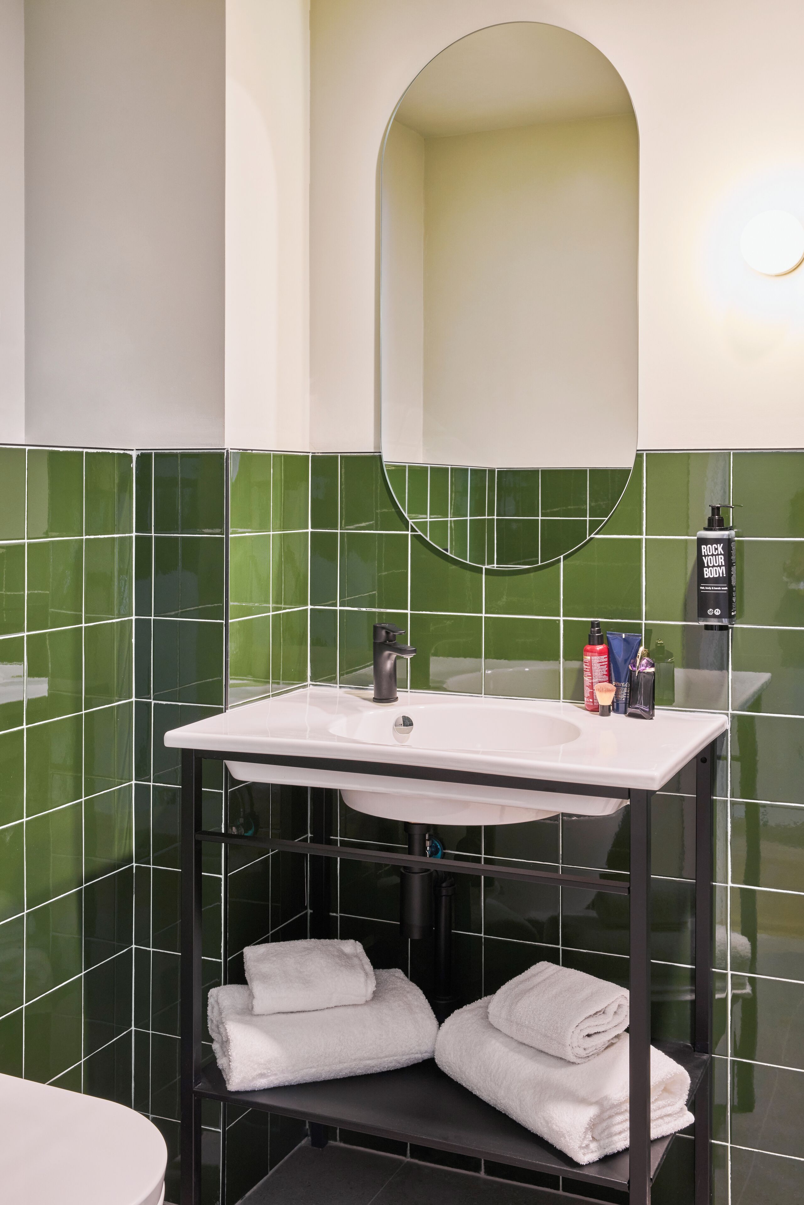 A bathroom with a white sink, green tiled walls, a mirror, and a shelf with folded towels.