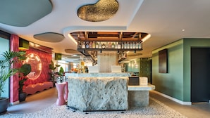 A modern lobby with a central stone reception desk, a staircase with a glass railing, and a variety of plants. - ibis Styles Madrid Airport Valdebebas (Madrid)