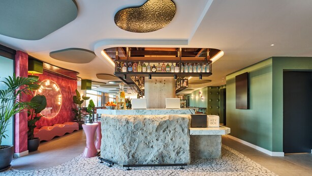 Eine moderne Lobby mit einer zentralen Empfangstheke aus Stein, einer Treppe mit Glastrennwand und verschiedenen Pflanzen.