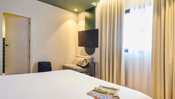 Habitación de hotel con una cama grande, una mesita de noche, una silla, un televisor y un ventanal con cortinas.