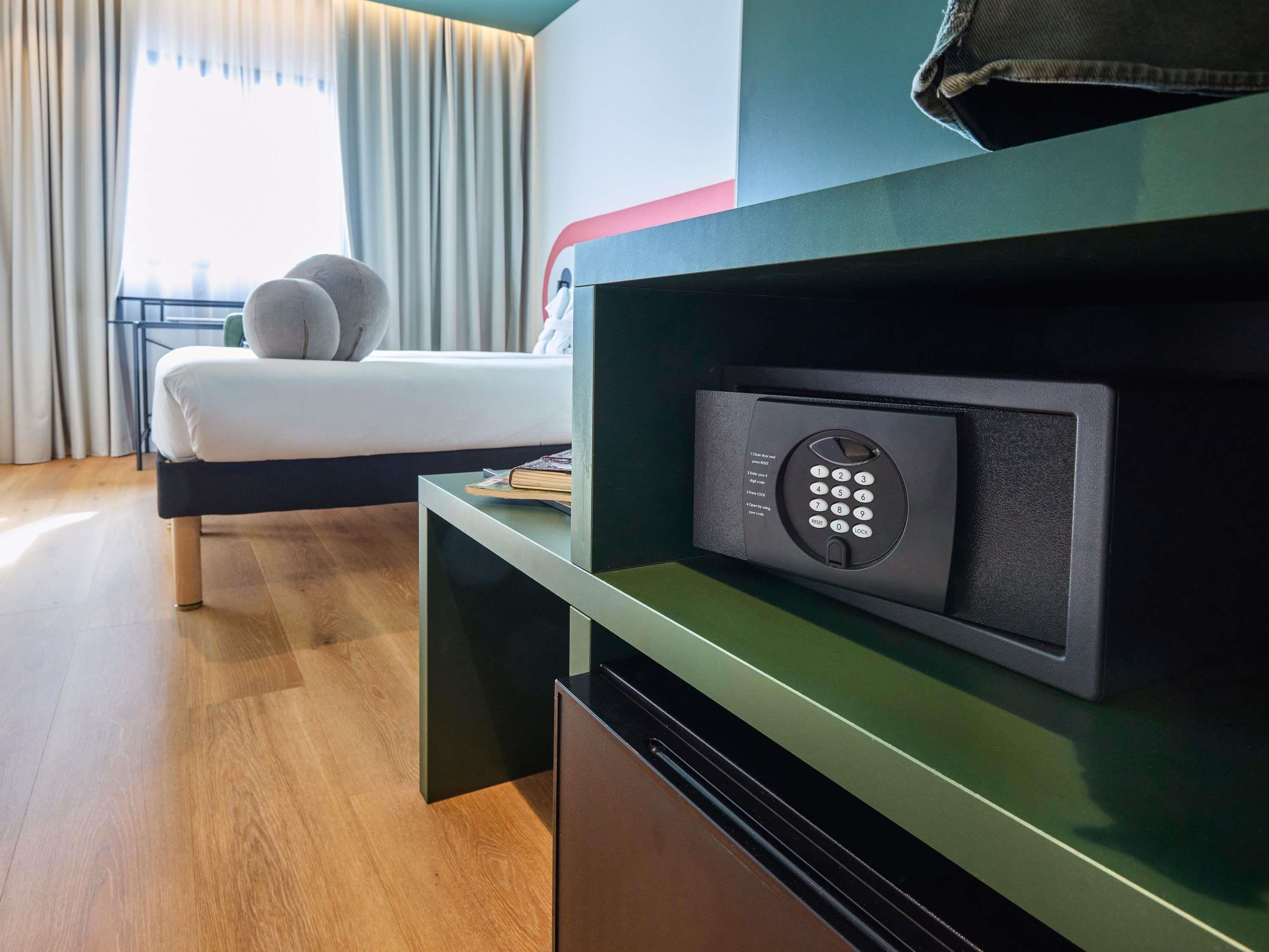 Ein modernes Hotelzimmer mit Bett, Nachttisch, fest eingebautem Safe und einem Fenster mit Vorhängen.