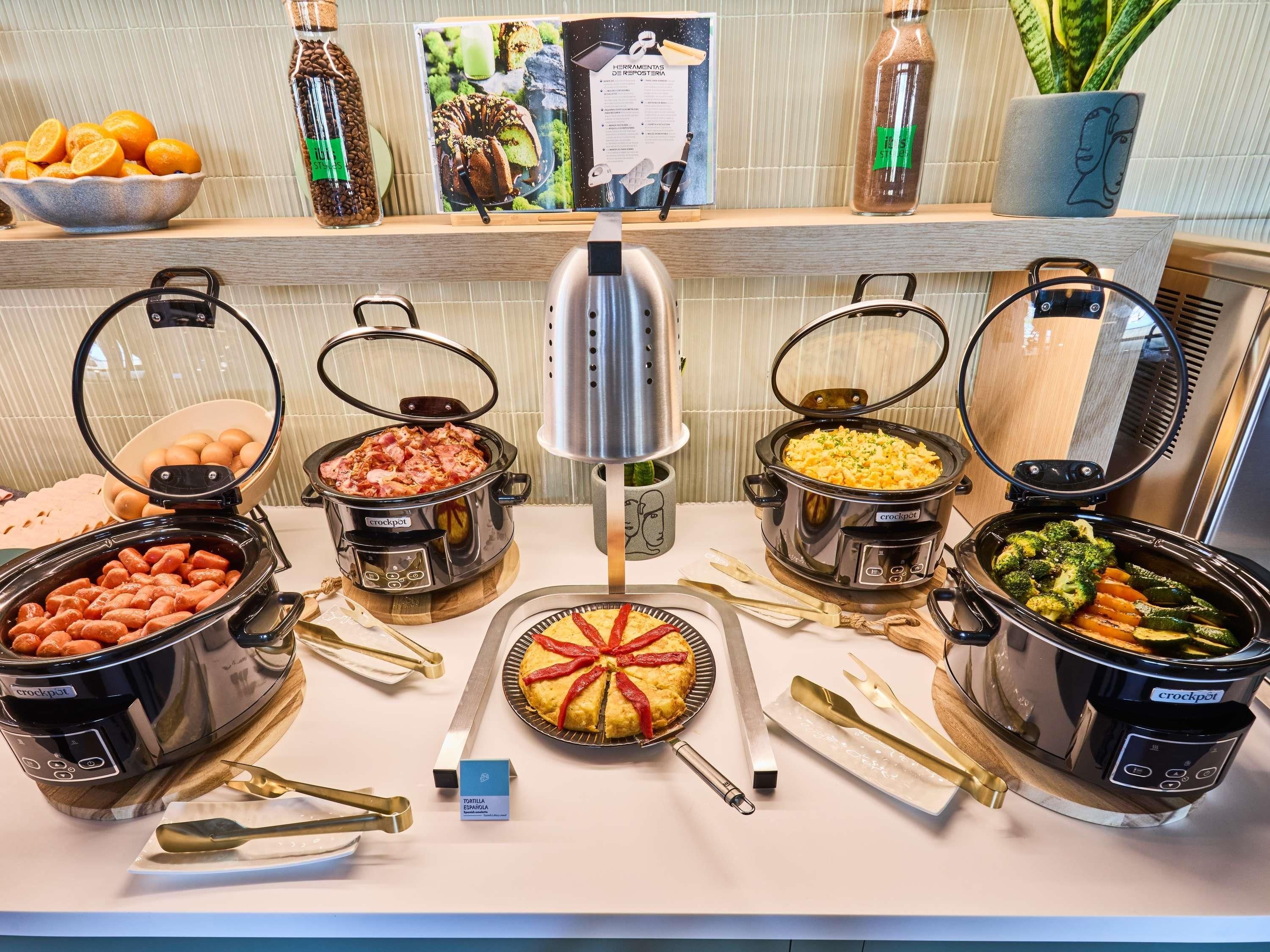 A buffet with various dishes in crockpots, a sign for Tortilla Española, and a display of oranges and coffee beans.
