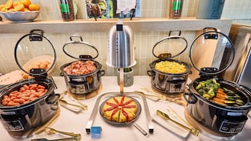 A buffet with various dishes in crockpots, a sign for Tortilla Española, and a display of oranges and coffee beans.