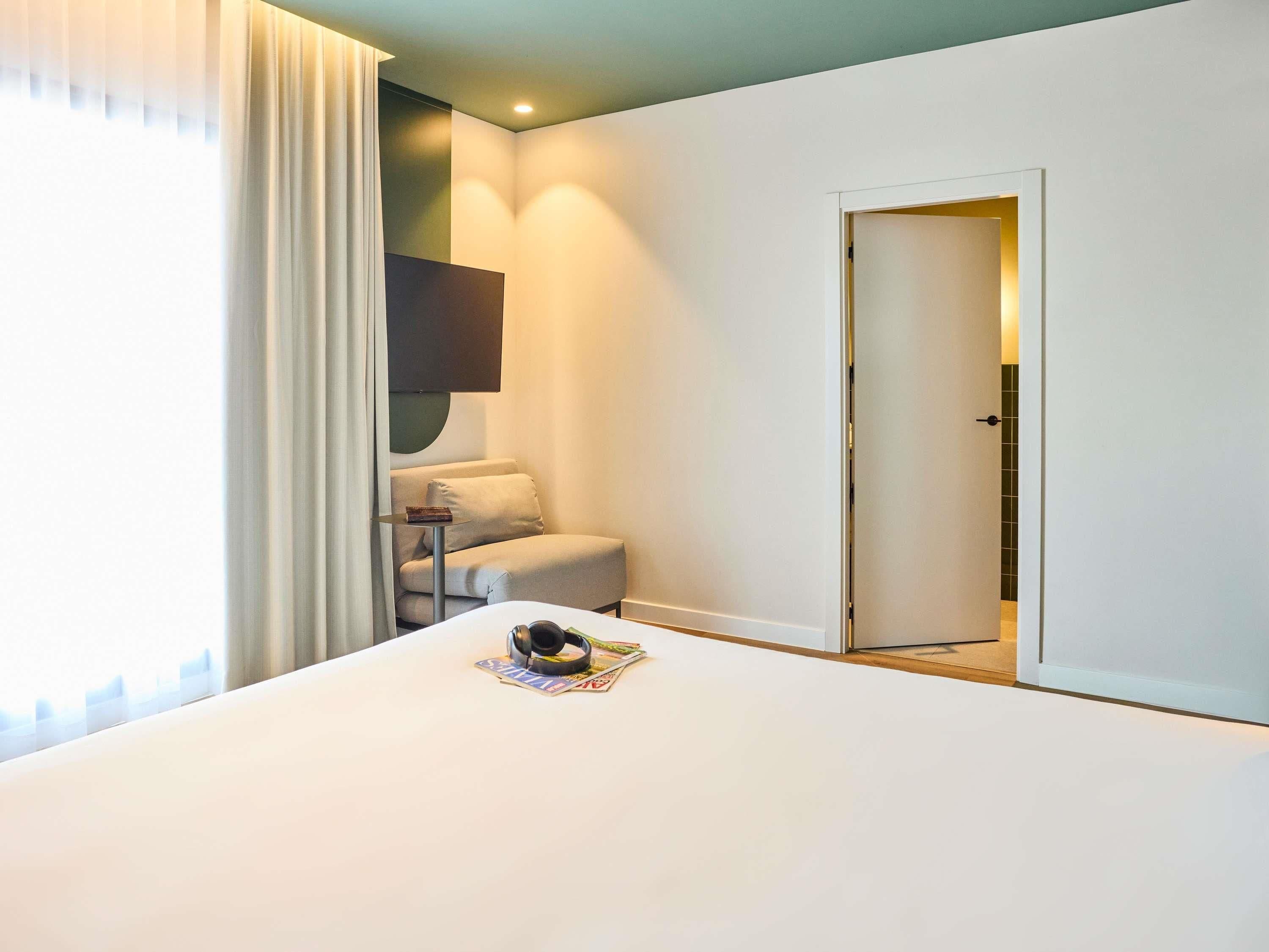 Habitación de hotel con una cama, una silla, una mesita, un televisor montado en la pared y una puerta que da a otra habitación.