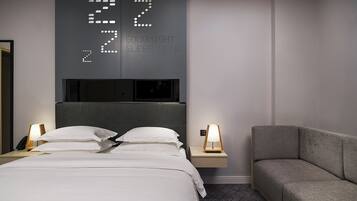 Une chambre d’hôtel moderne avec un grand lit, des tables de chevet avec des lampes, un canapé gris et une horloge numérique fixée au mur qui affiche l’heure.