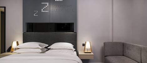 Una camera d'hotel moderna con un letto grande, comodini con lampade, un divano grigio e una sveglia digitale a parete che indica l'ora.