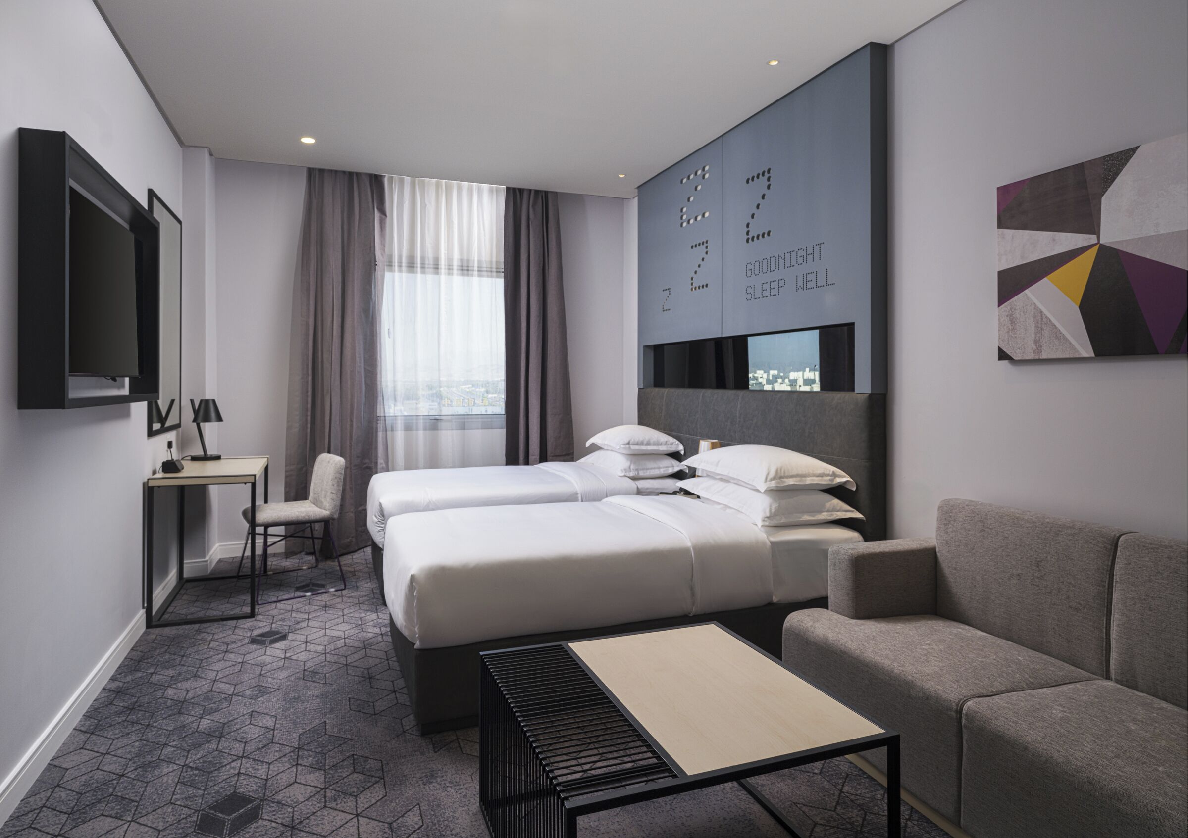 A modern hotel room with a bed, two bedside tables, a sofa, a TV, and a city view.