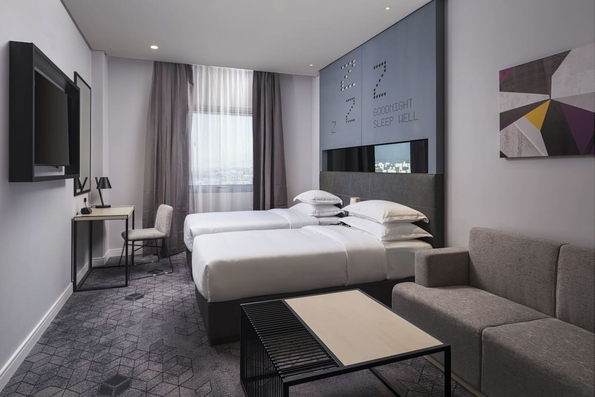 A modern hotel room with a bed, two bedside tables, a sofa, a TV, and a city view.