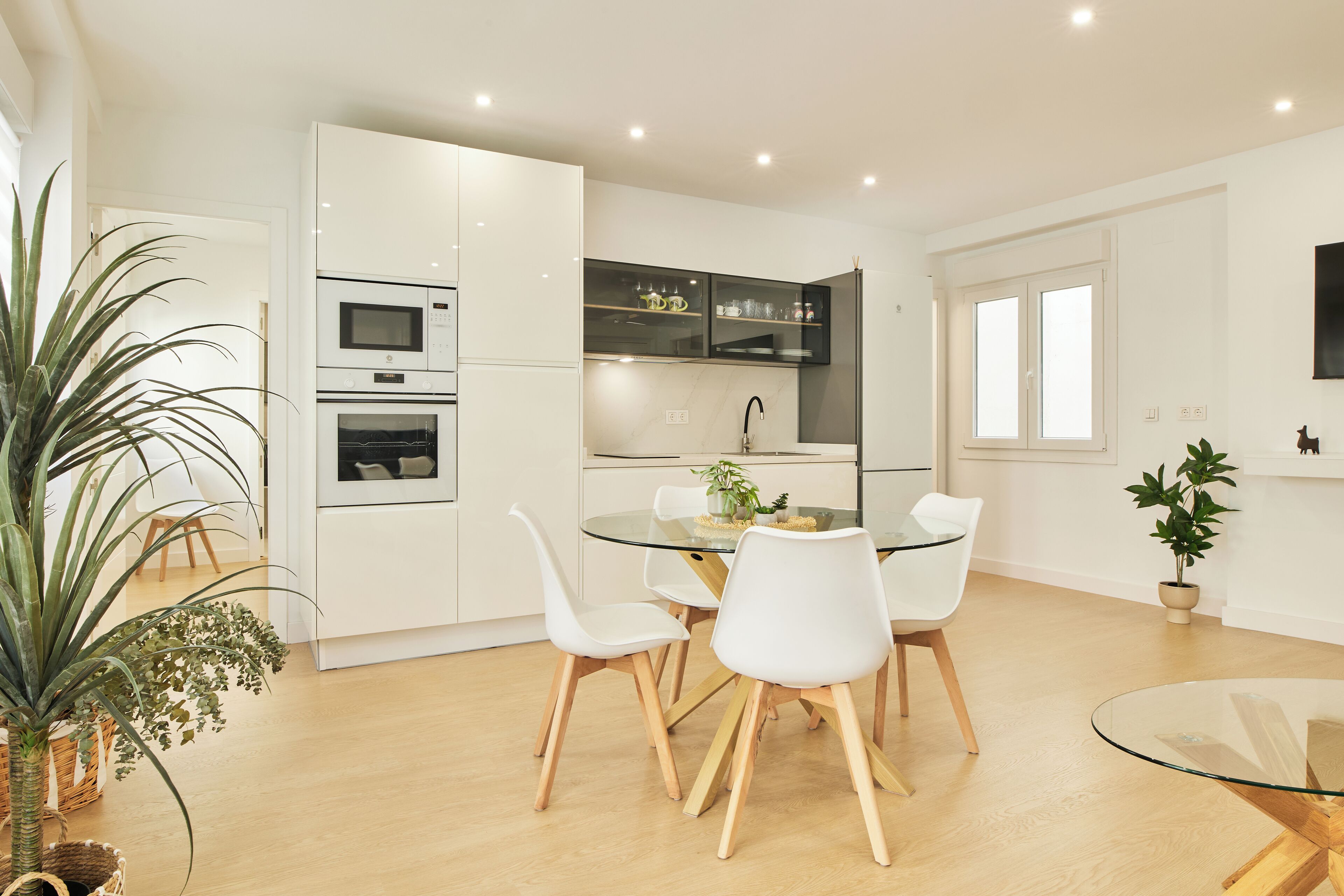 Une cuisine moderne avec des meubles blancs, un four encastré et un four à micro-ondes. On y trouve également un coin repas avec une table en verre et des chaises blanches.