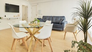 A modern living room with a glass dining table, white chairs, a grey sofa, and a large potted plant.