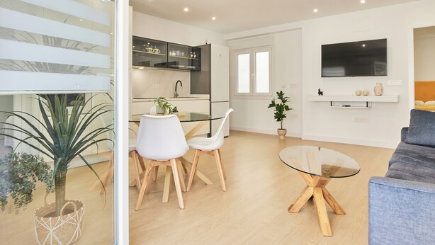A modern living room with a glass dining table, white chairs, a grey sofa, and a TV mounted on the wall.