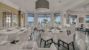 Un restaurant où les tables sont dressées pour le service, avec de grandes fenêtres offrant une vue agréable et un décor intérieur moderne.