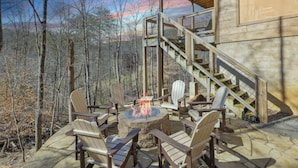 Een houten blokhut met een vuurkuil van steen, omringd door Adirondack stoelen, een trap en uitzicht op een bebost landschap.