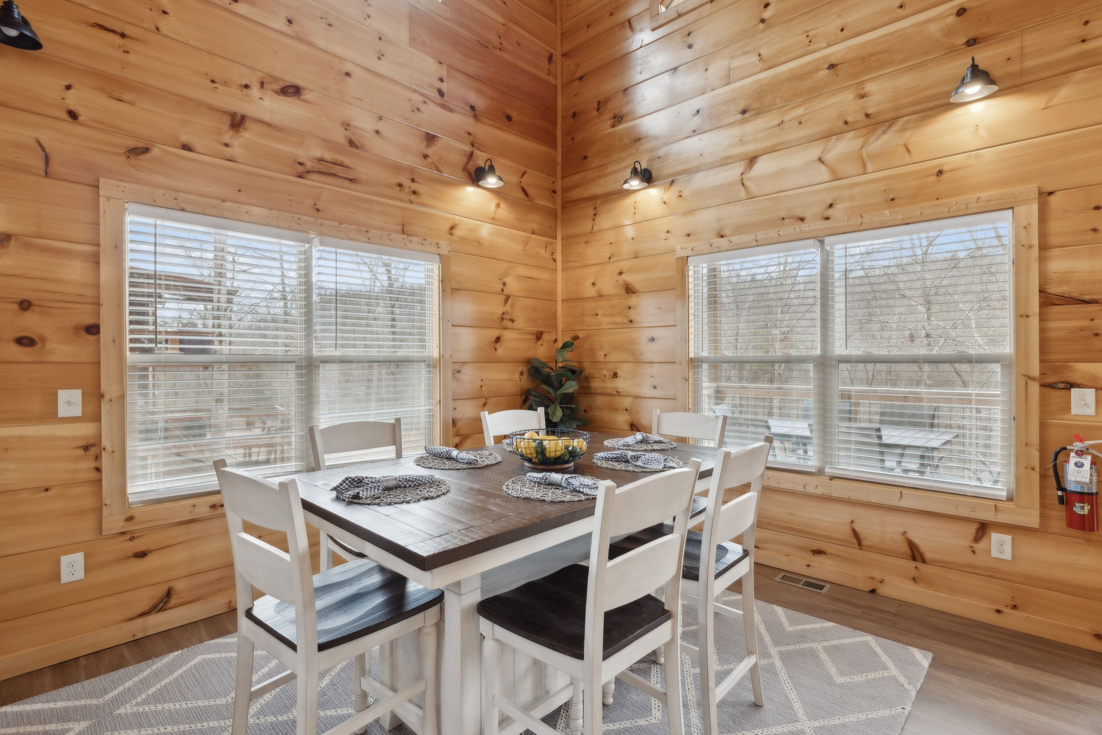 Comedor con paneles de madera, una mesa dispuesta para cuatro comensales, dos luces fijadas a la pared y ventanas amplias con persianas.